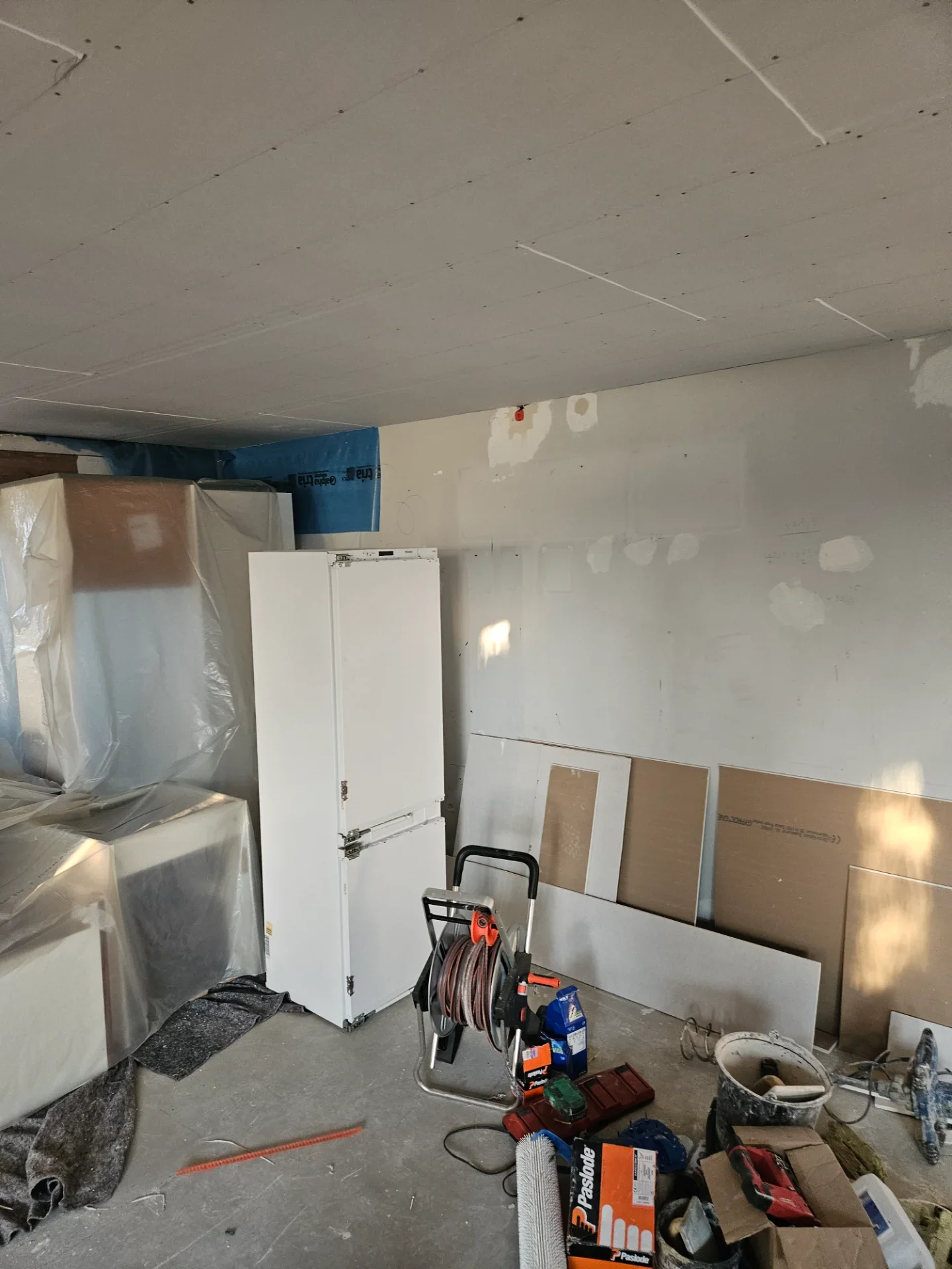 Billede af et køkken under installation med køleskab, værktøj og byggematerialer.