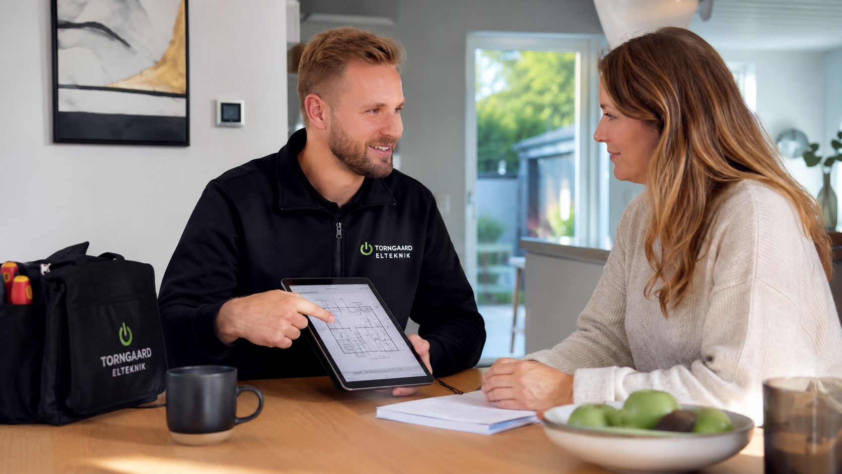 En professionel elinstallatør gennemgår plantegninger på en tablet sammen med en kunde ved et spisebord.